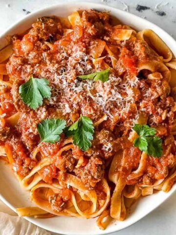 a plate of slow cooker lamb ragu over pappardelle pasta surrounded by a block of parmesan, a white linen, and torn baguette.