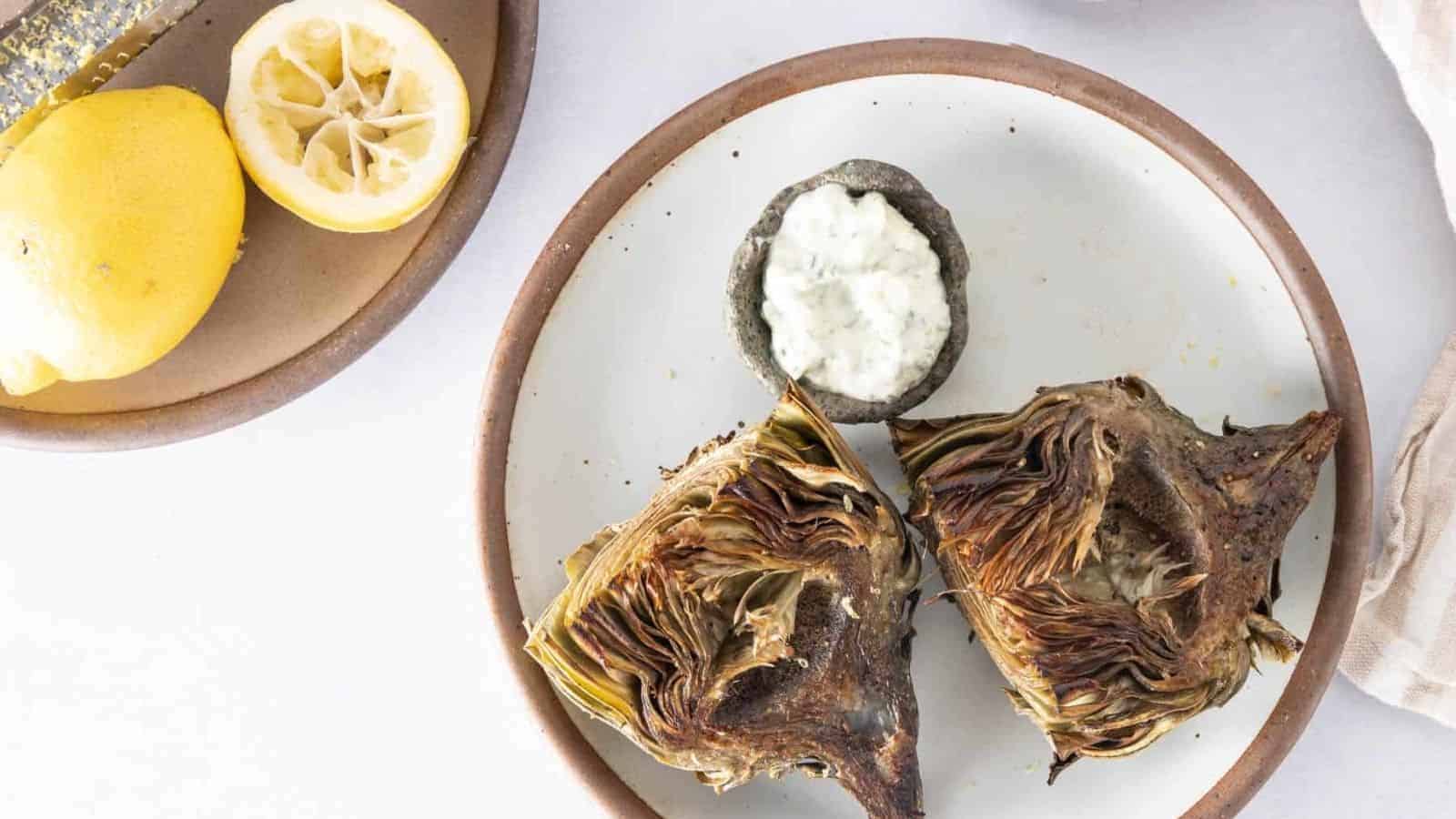 A white plate with two grilled artichoke halves and a small stone bowl of dipping sauce.