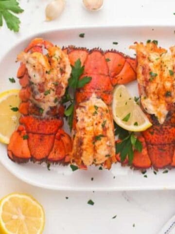 Top view of air fryer lobster tails on a plate with lemon slices and greens as garnishes.
