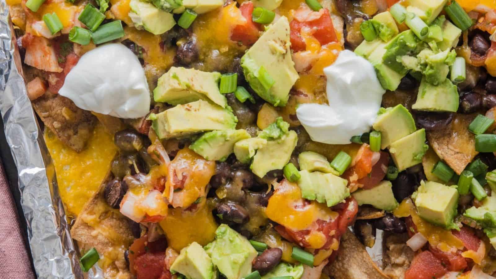 A close-up imag of loaded nachos topped with melted cheese, avocado chunks, black beans, diced tomatoes, green onions, and dollops of sour cream.