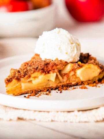 A close-up photo of a sliced apple pie with graham cracker crust on a white plate, topped with a scoop of vanilla ice cream. 