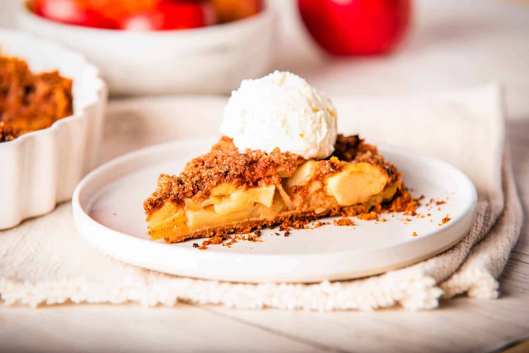 A close-up photo of a sliced apple pie with graham cracker crust on a white plate, topped with a scoop of vanilla ice cream. 