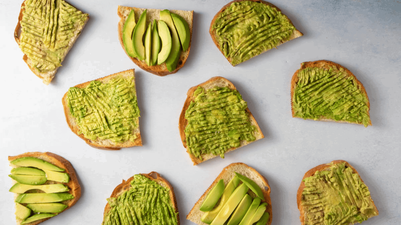 An overhead image of various slices of bread, some topped with avocado slices, others toasted with fork markings.