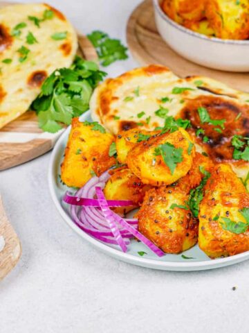 Horizontal shot of Bombay Potatoes on a plate with naan, with extra naan and red onions on the side.