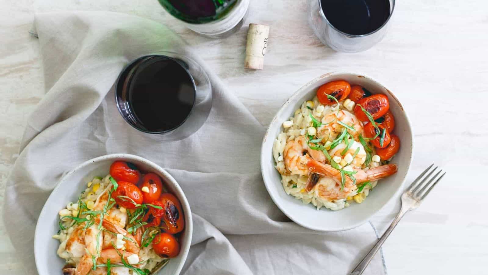 Overhead view of two bowls of brown butter shrimp with parmesan basil corn orzo, with two glasses of wine and a wine bottle in the background.