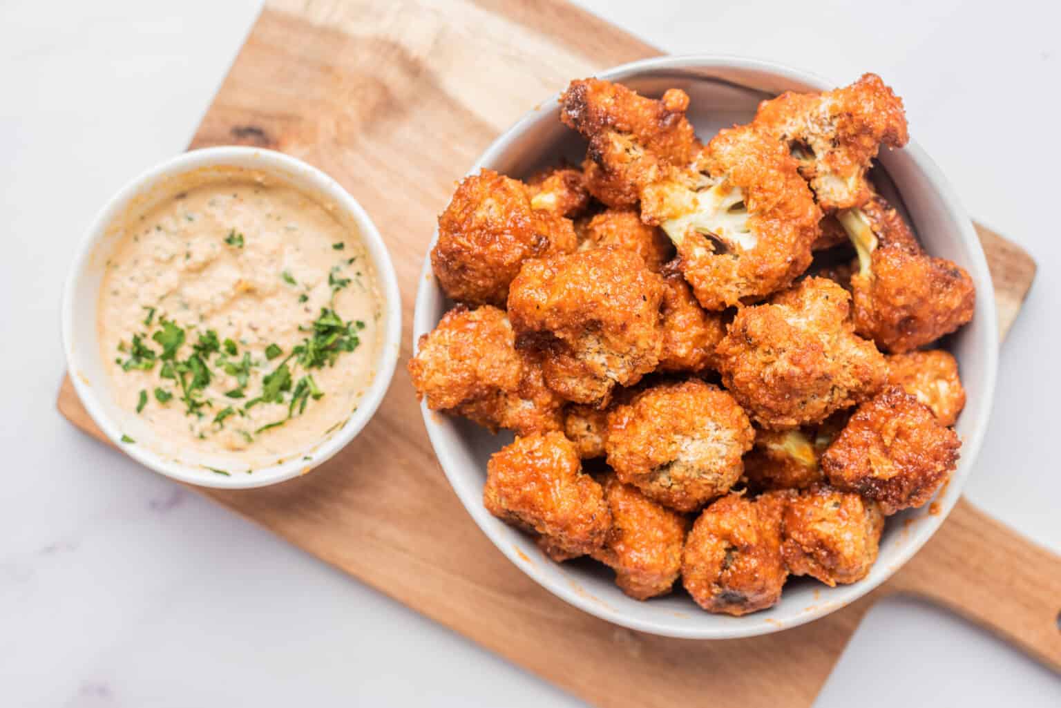 Air fryer vegan buffalo cauliflower bites in a bowl with dip nearby, placed under a wooden board.
