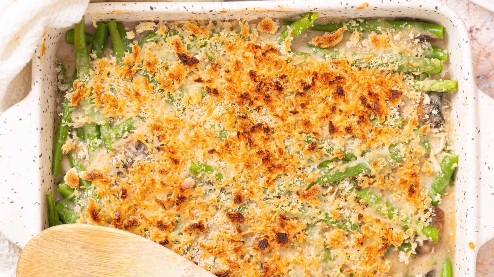 An image of a white plate with a serving of cheesy green bean casserole with a fork on the side, and the casserole behind it.