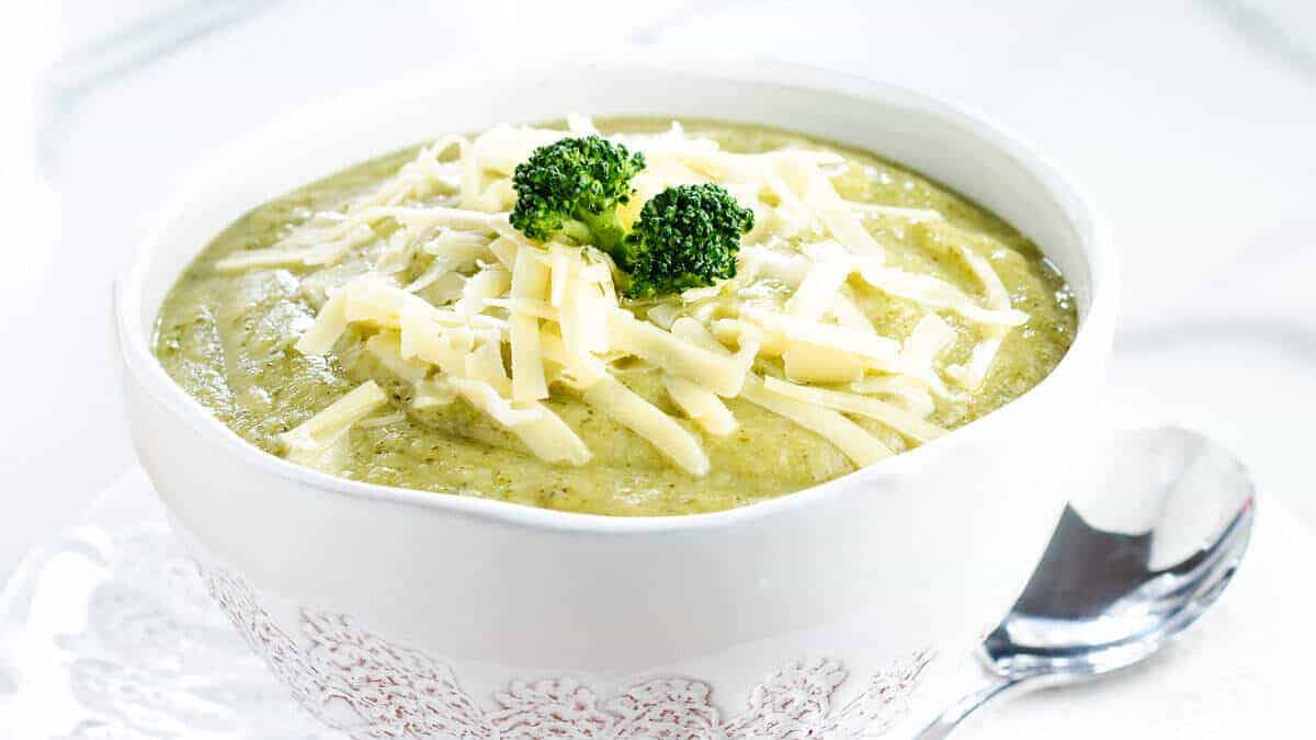 A close up image of broccoli soup in a white bowl topped with shredded cheese.