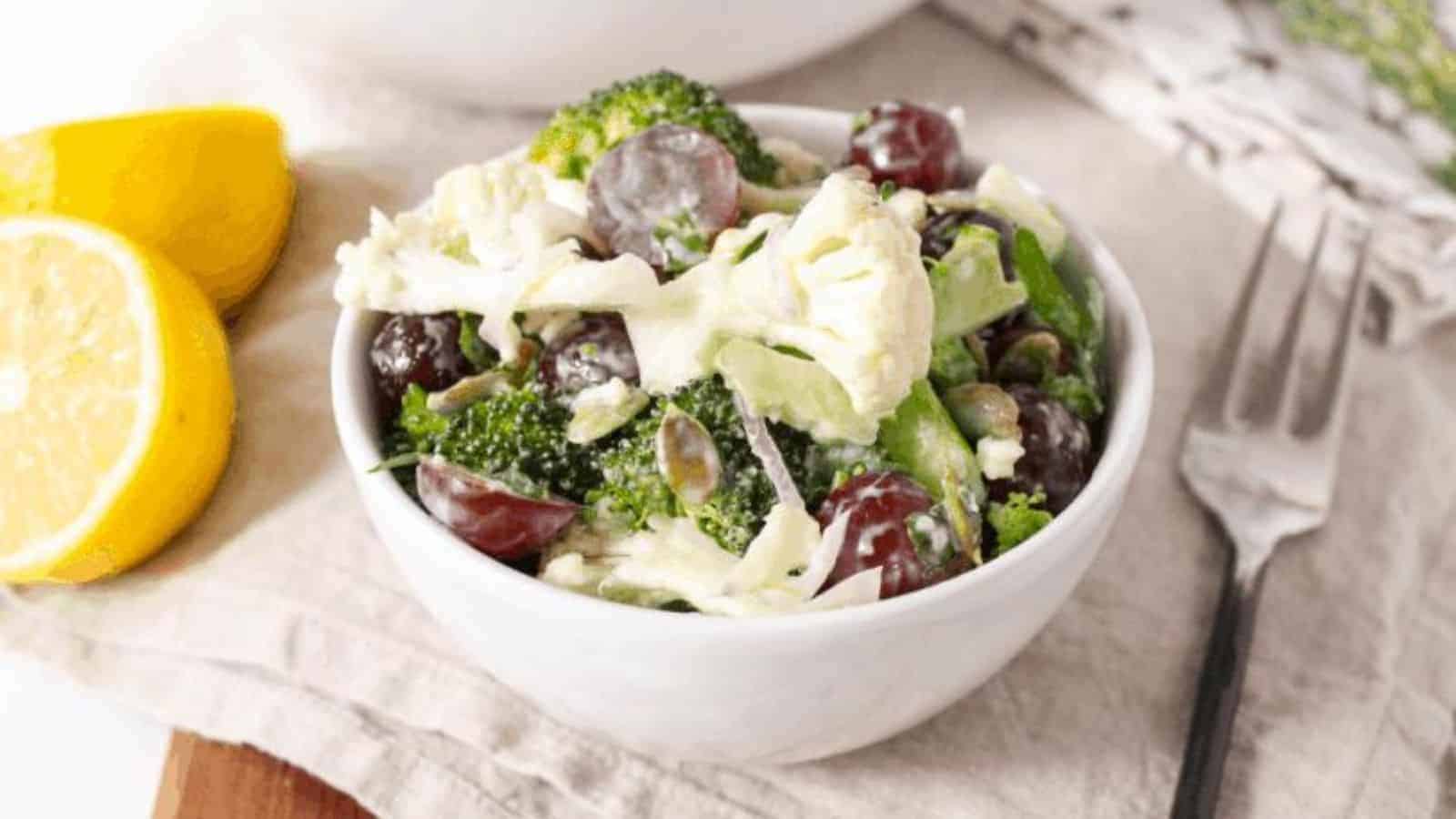 A small white bowl with broccoli, cauliflower, grapes, and pumpkin seeds tossed in a yogurt dressing.