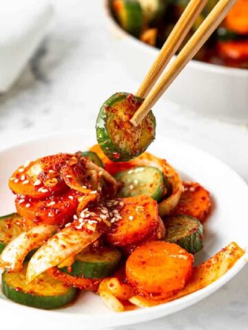 Cucumber kimchi on a white plate with a pair of chopsticks lifting a slice of cucumber.