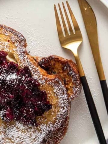 A close-up of French toast topped with berry compote, accompanied by utensils and a cup of coffee.