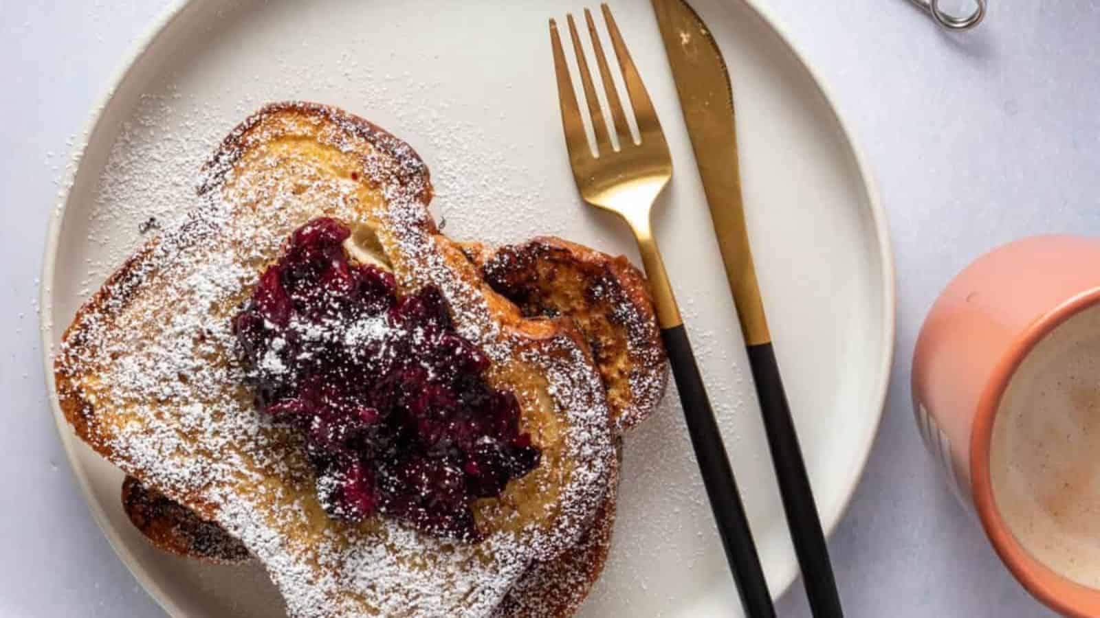 A close-up of French toast topped with berry compote, accompanied by utensils and a cup of coffee.
