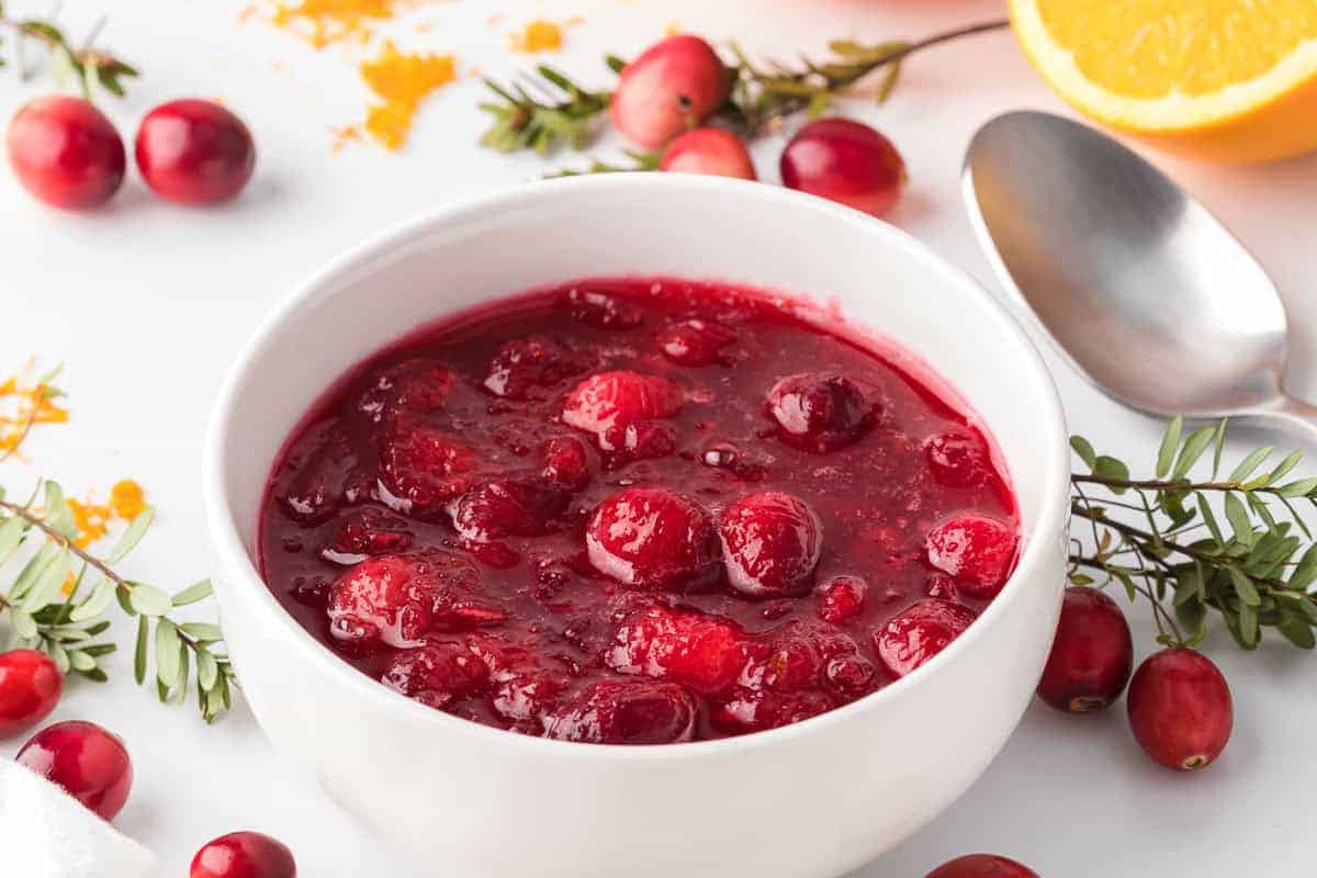 A close up photo of fresh cranberry sauce placed in a white bowl with spoon, oranges and fresh cranberries in the background.