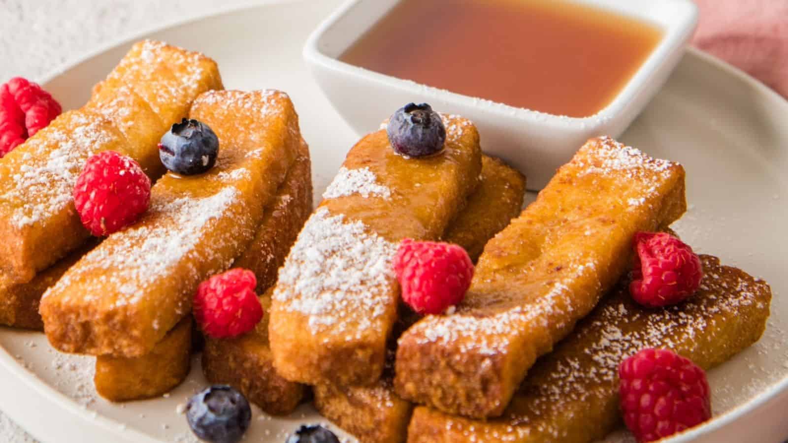 A plate of golden brown French toast sticks dusted with powdered sugar, garnished with raspberries and blueberries.