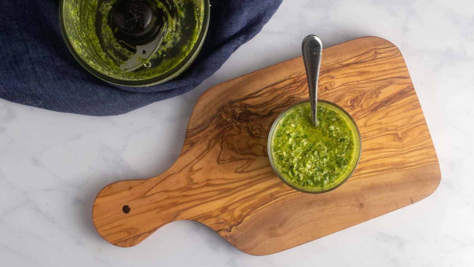 An overhead image of green goddess salad dressing in a glass jar, with a spoon inside, placed on a wooden board beside a blender.