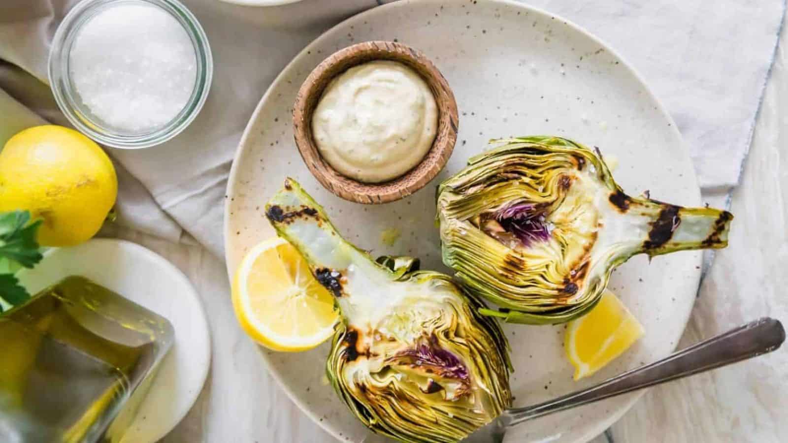 Overhead image of grilled artichokes with a dip and lemon slices on a plte.