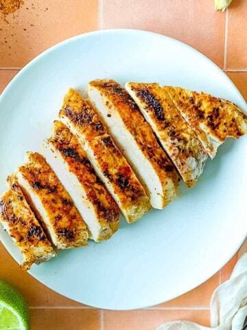 Sliced Tajín Grilled Chicken sits on a white plate surrounded by Tajín seasoning, a sliced lime, a white linen, and a small glass of beer.
