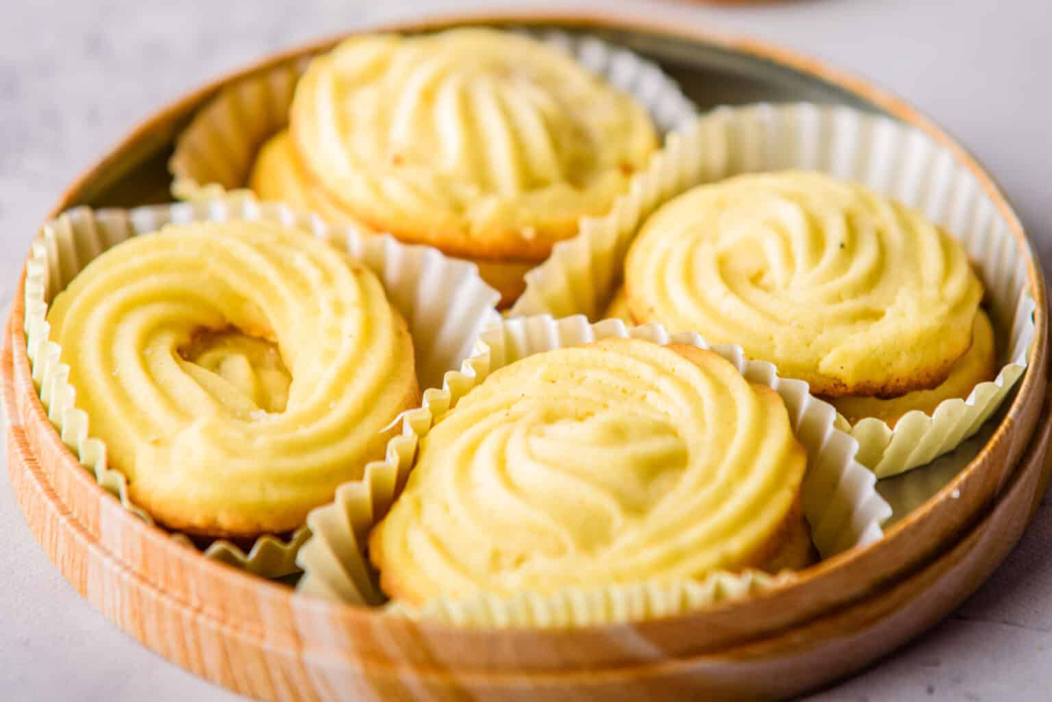 Four swirled butter cookies, each nestled in a white paper liner, arranged on a wooden tray.