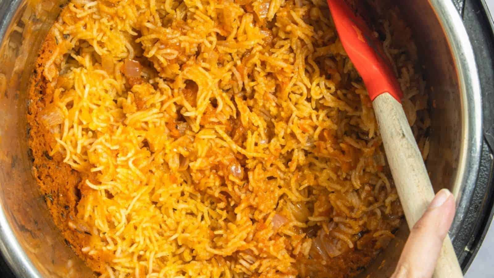 A close-up of a pot with cooked jollof rice with a person's hand holding a red spatula.