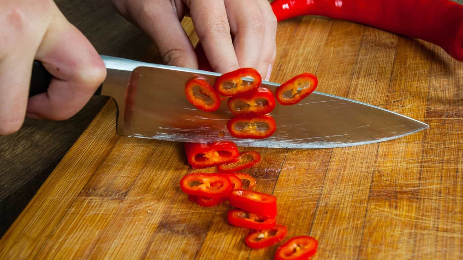 Hands chopping chili pepper on wooden board.