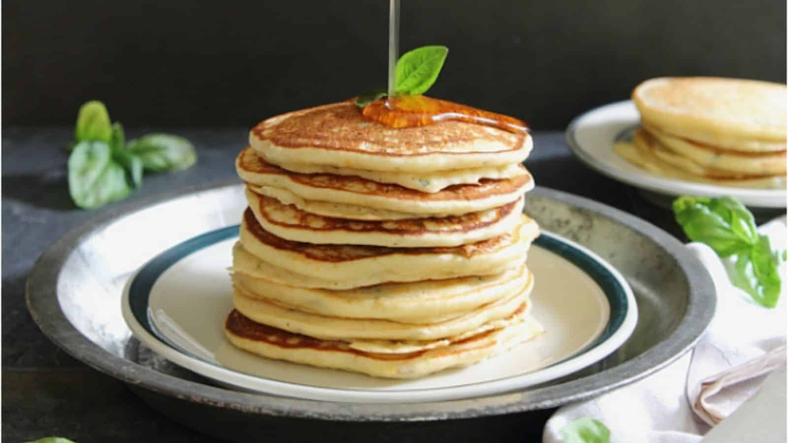 A plate of stacked orange basil ricotta pancakes with syrup pouring onto it.