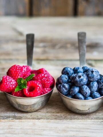 Metal measuring cups filled with blackberries, raspberries, blueberries, and strawberries are arranged in a row on a wooden surface.