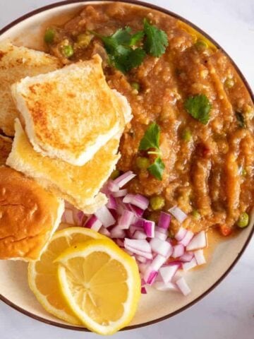 A top-down view of a plate with pav rolls, bhaji masala, red onions, lemon wedges, and garnishes on the side.