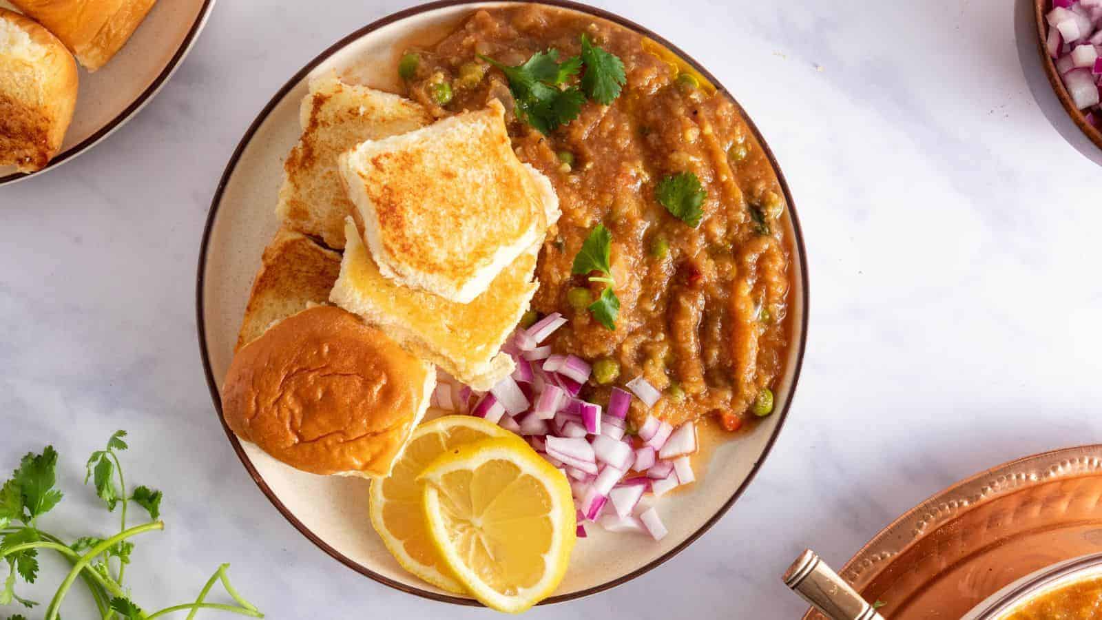 A top-down view of a plate with pav rolls, bhaji masala, red onions, lemon wedges, and garnishes on the side.