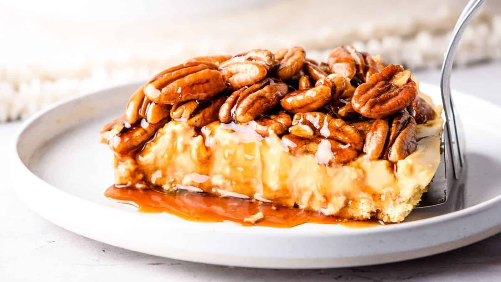 A close up view of pecan pie cheesecake slice on a white plate, with a fork on the side.