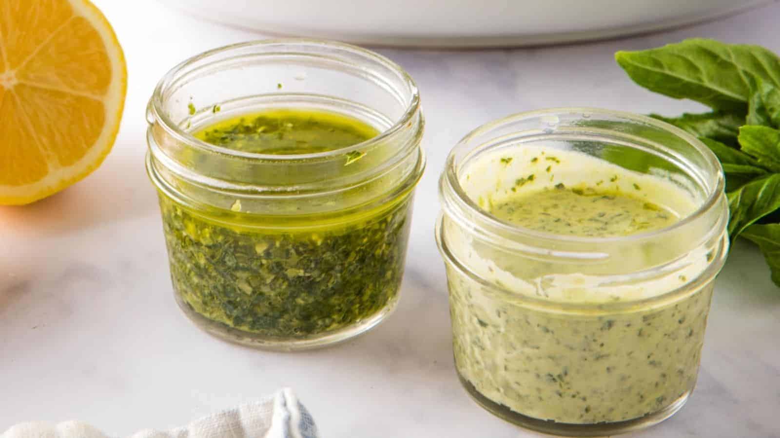 Two small glass jars filled with pesto salad dressings, one vinaigrette and one creamy, on a white marble surface.