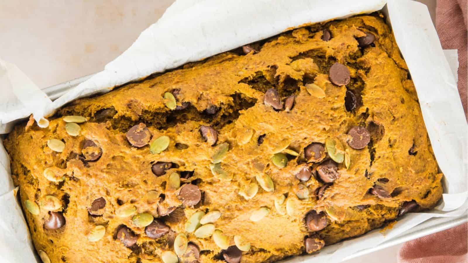 An overhead view of a freshly baked loaf of pumpkin bread with chocolate chips and pumpkin seeds sits in a parchment-lined loaf pan.