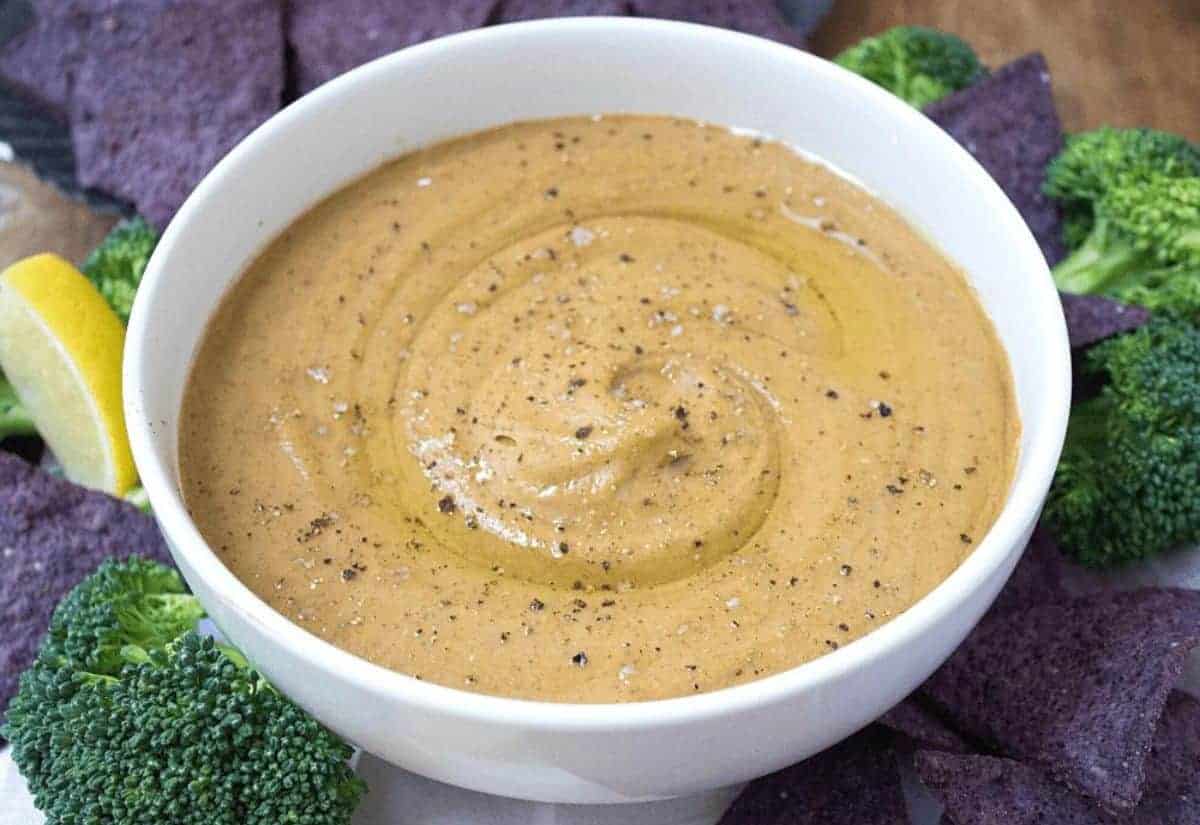 A white bowl of creamy pumpkin dip with olive oil swirled into it, surrounded by broccoli and purple tortilla chips.