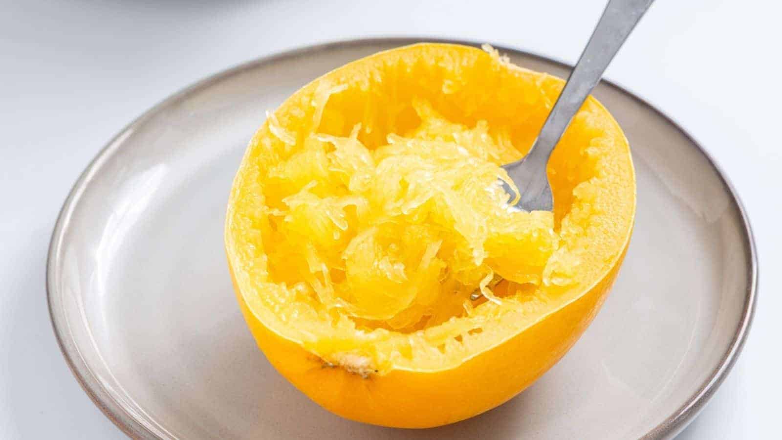 A halved spaghetti squash on a plate with a fork scooping out the stringy, cooked flesh.