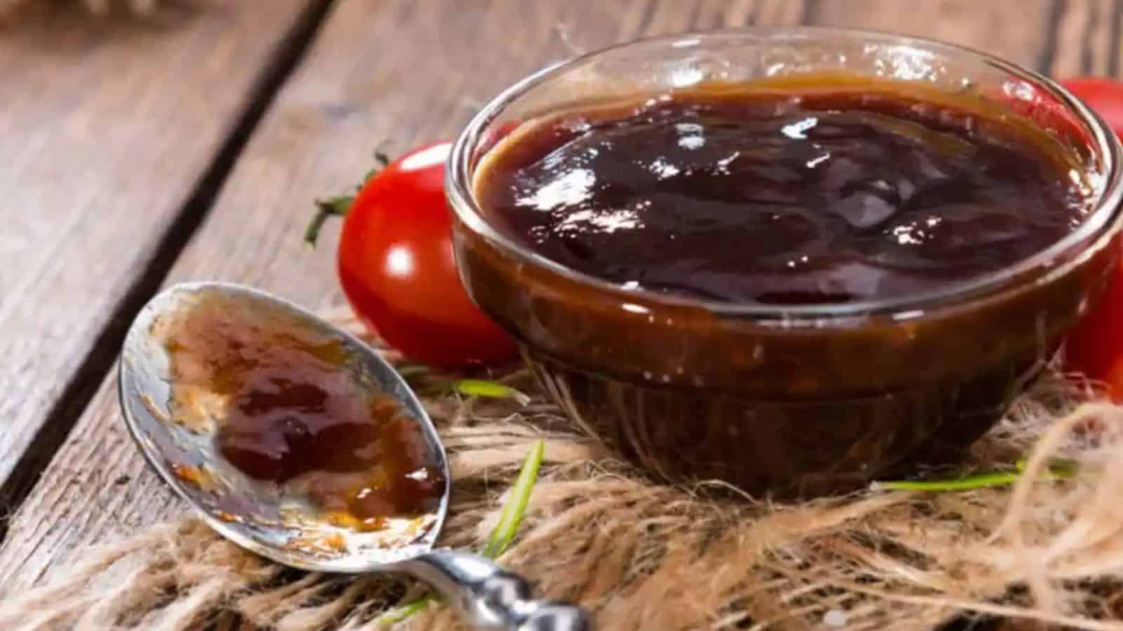 A small glass bowl filled with barbecue sauce sitting beside a spoon with barbecue sauce.