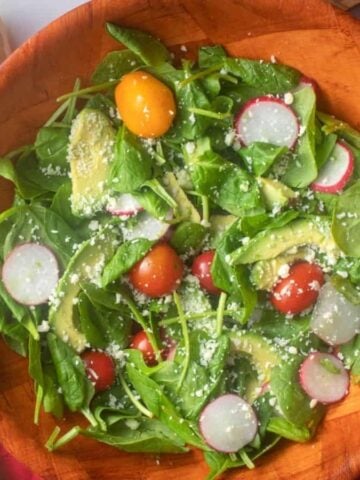 An overhead image of spinach avocado salad in a wooden bowl with lemon in the background.