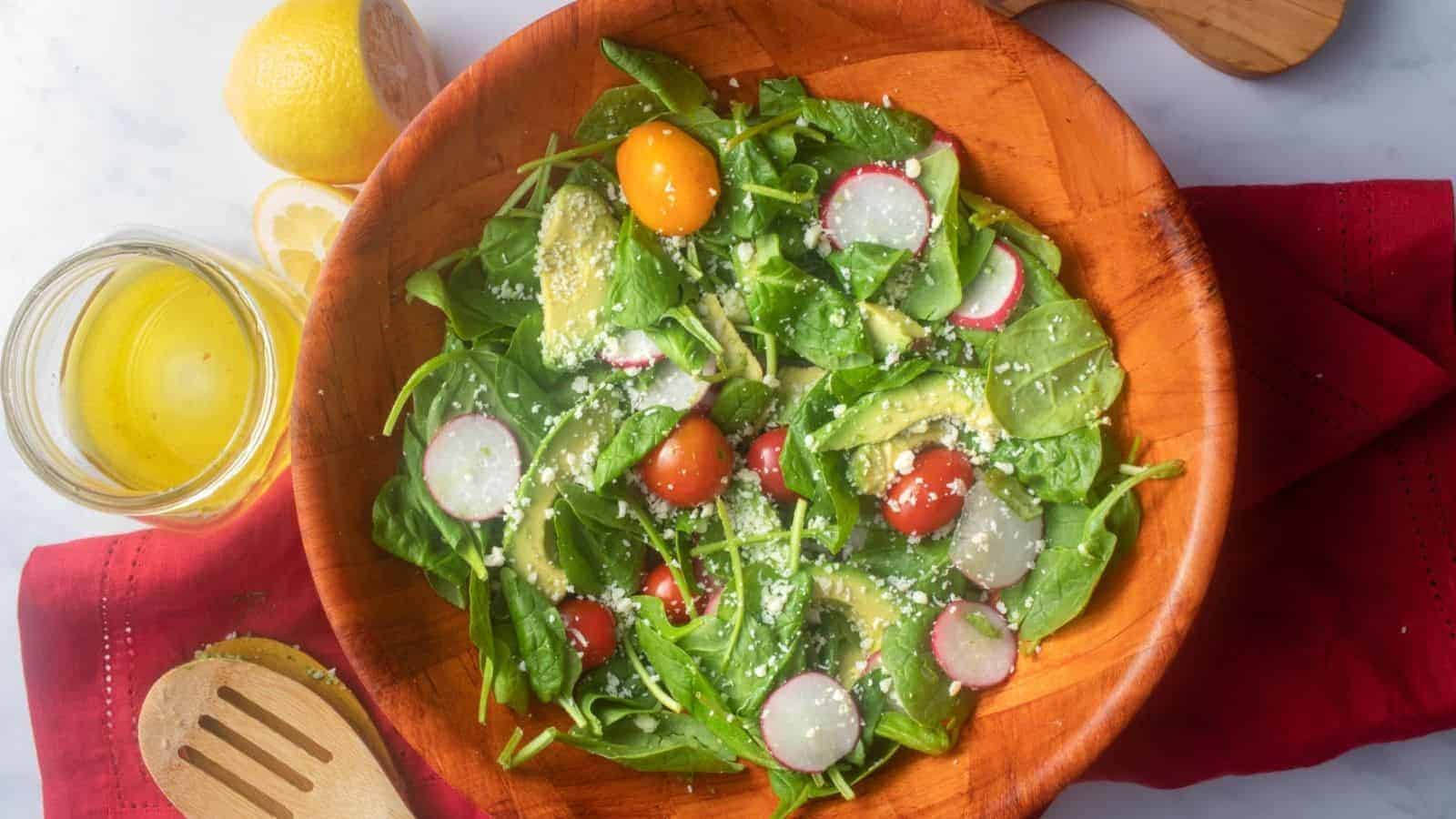 An overhead image of spinach avocado salad in a wooden bowl with lemon in the background.