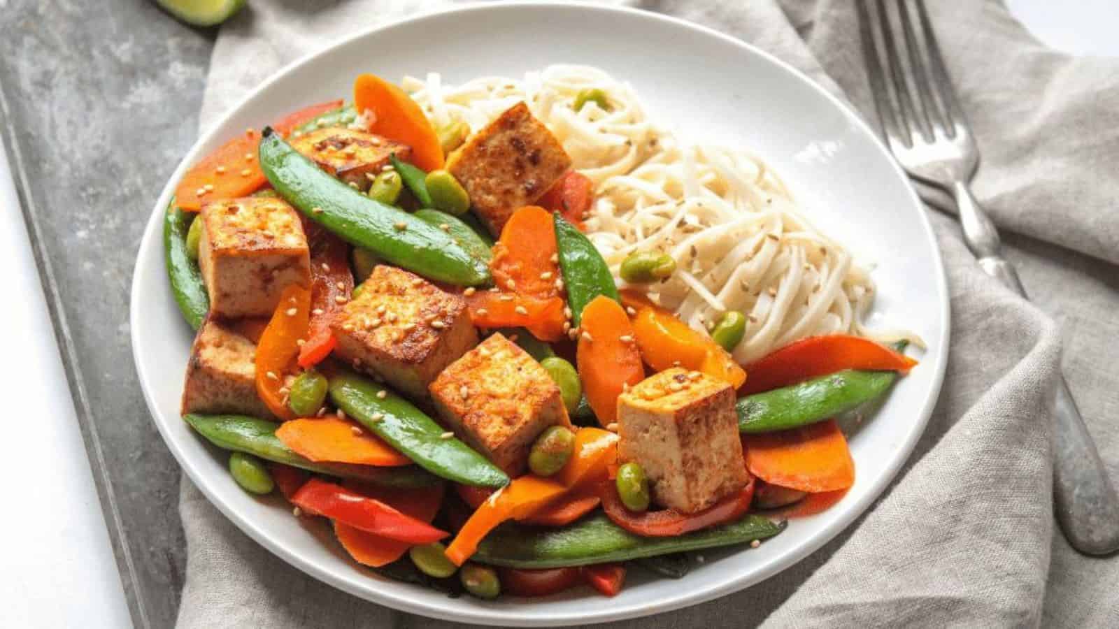 Seared tofu with roasted carrots, edamame, and bell peppers next to rice noodles on a white plate.