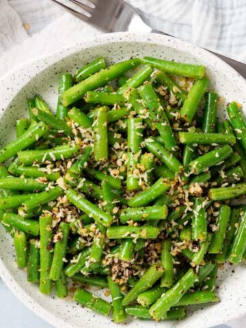 A top-down view of stir-fried green beans with coconut on a white plate, accompanied by a white napkin and fork.