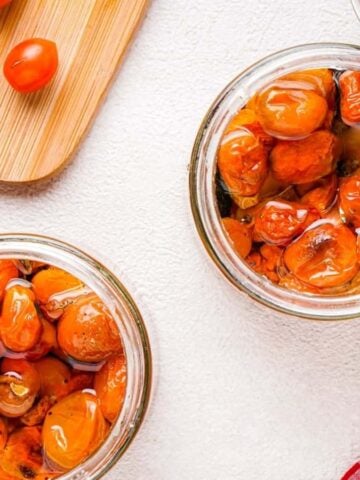 Two glass jars of cherry tomatoes and garlic confit on a white surface.