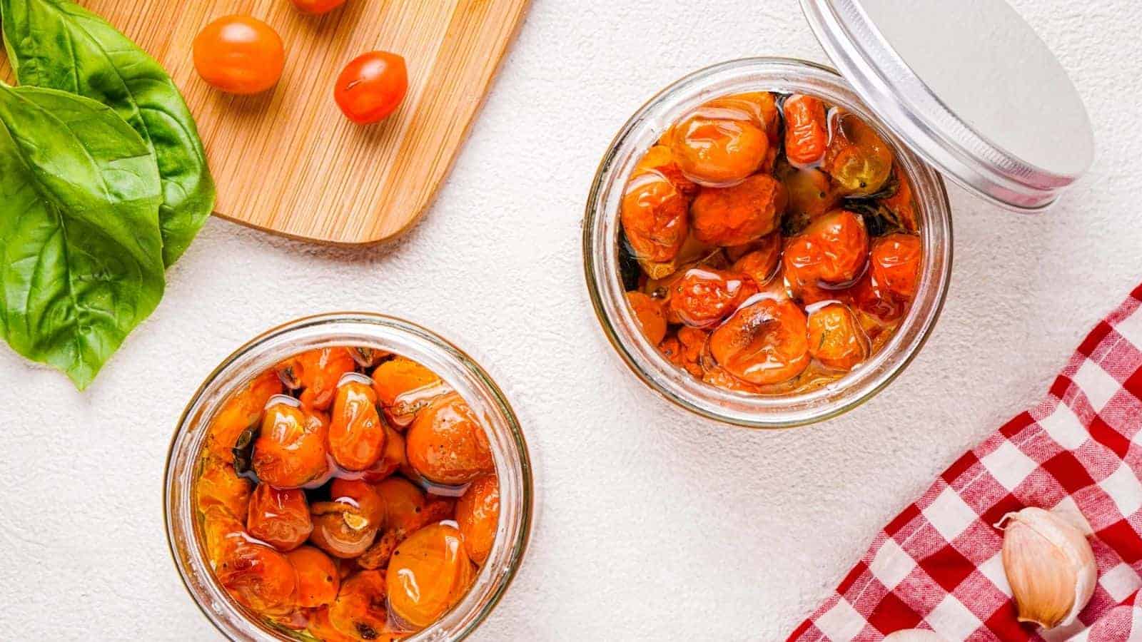 Two glass jars of cherry tomatoes and garlic confit on a white surface.