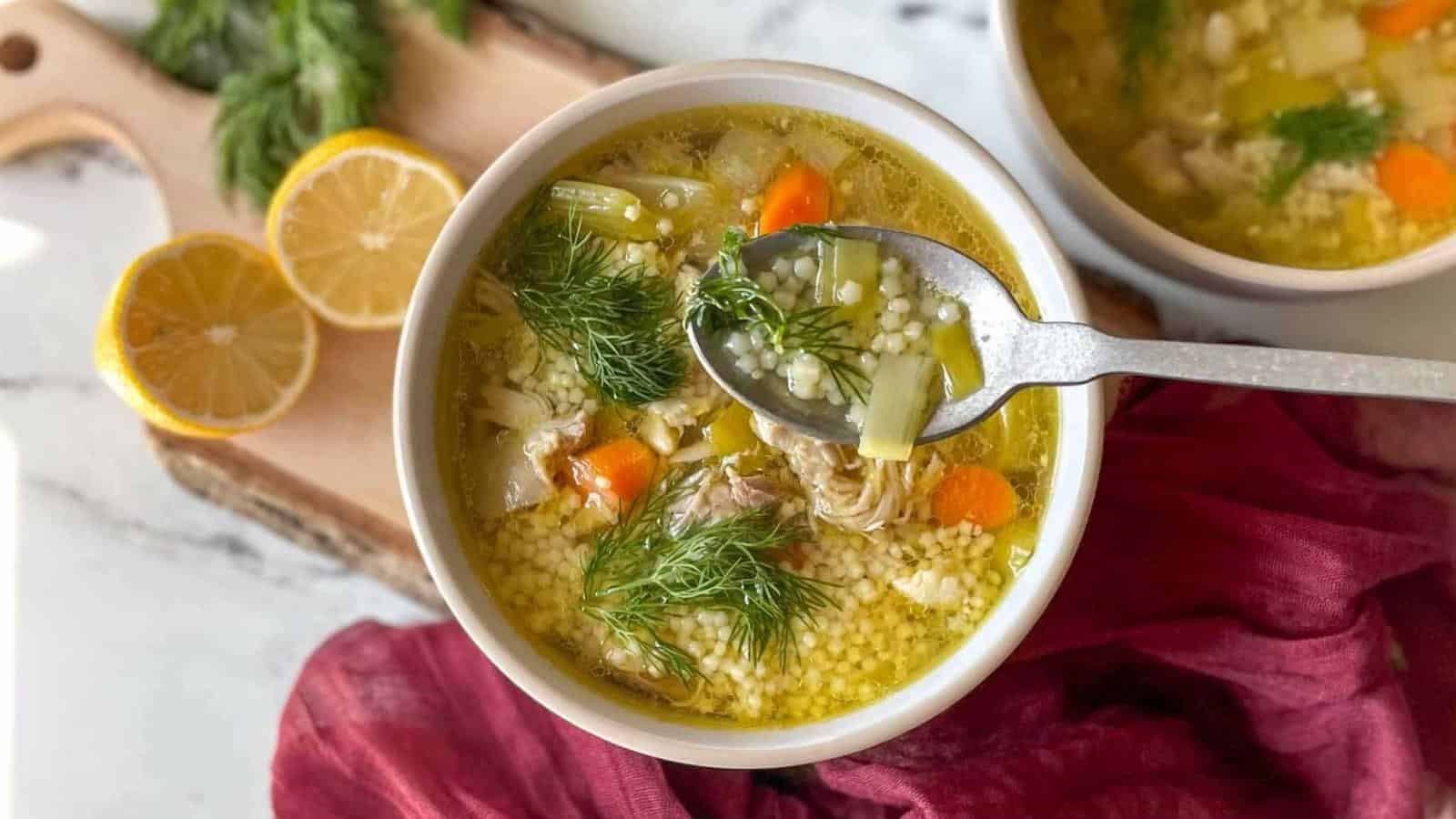 A bowl of chicken pastina soup sits on a rustic wooden cutting board with lemons and dill in the background.