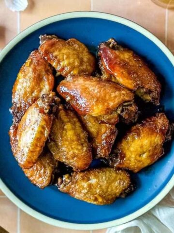 Soy garlic wings on a blue plate surrounded by a white linen, a glass of bear, soy ginger sauce, and cloves of garlic.