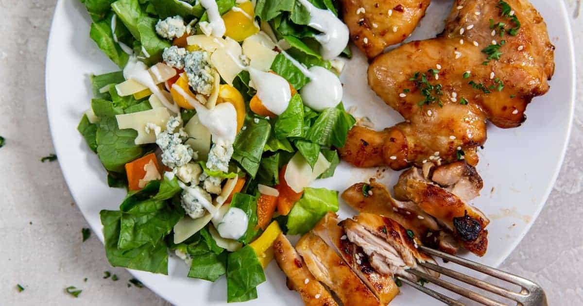 Air Fryer Teriyaki Chicken Thighs Paleo on a white plate with parsley, sesame seeds, lettuce with bell peppers, bleu cheese, parmesan and a fork.