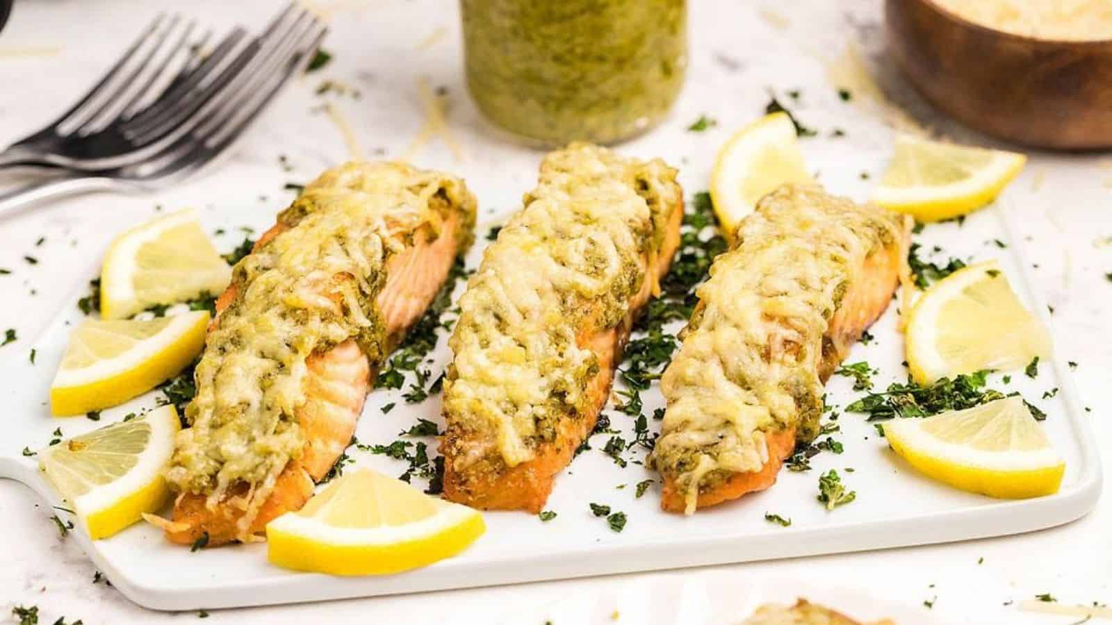 Close-up image of three parmesan pesto crusted salmon fillets on a plate garnished with chopped greens and lemon slices.