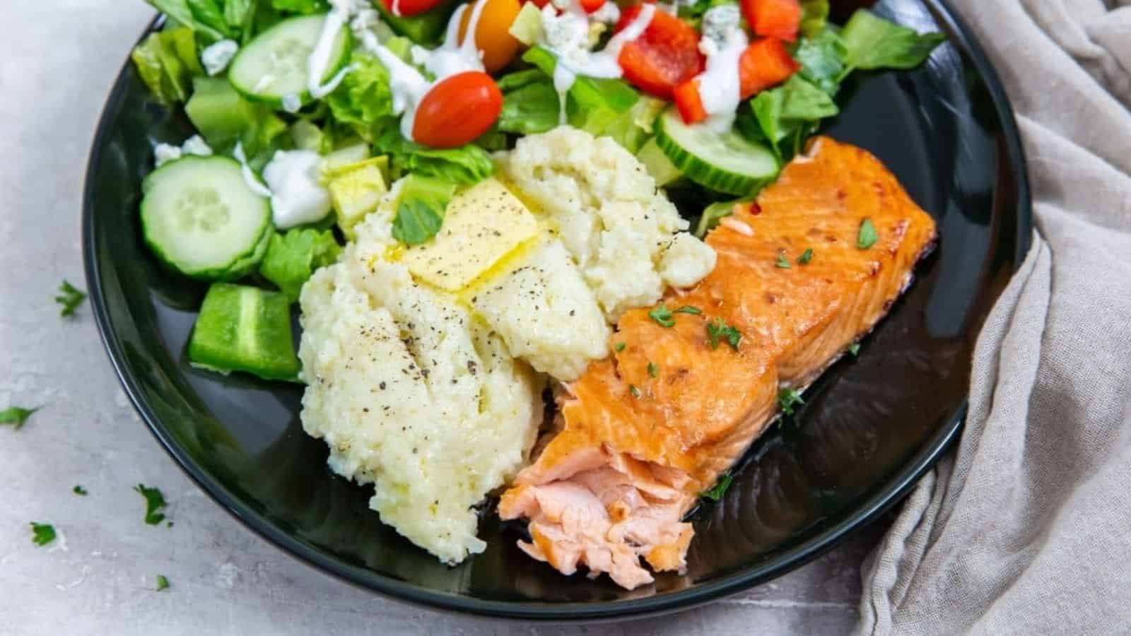 Air Fryer Teriyaki Salmon with rice, butter, parsley, black pepper, and a salad on a black plate.