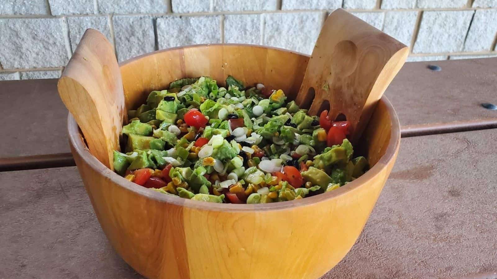 Image shows a wooden bowl containing avocado tomato salad shown from overhead and sitting on a picnnic table.