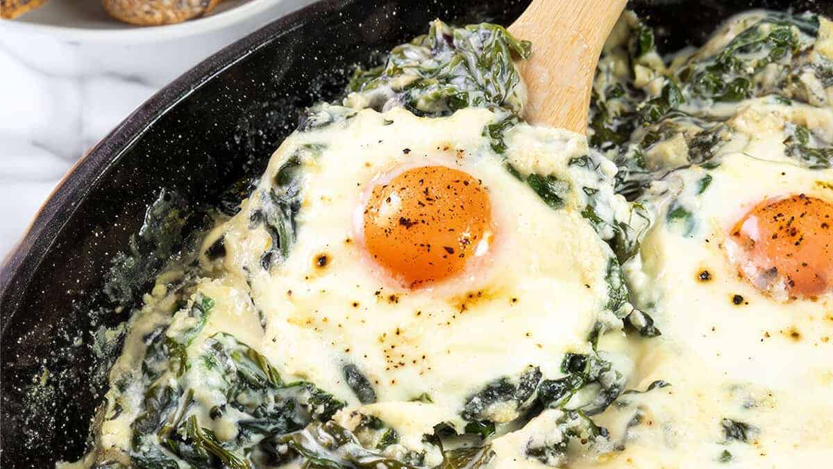 Baked Eggs Florentine up close. It showcases eggs baked in a skillet, with one egg's yolk front and center, bright and slightly runny. The eggs are embedded in a rich mix of melted cheese and creamy spinach.