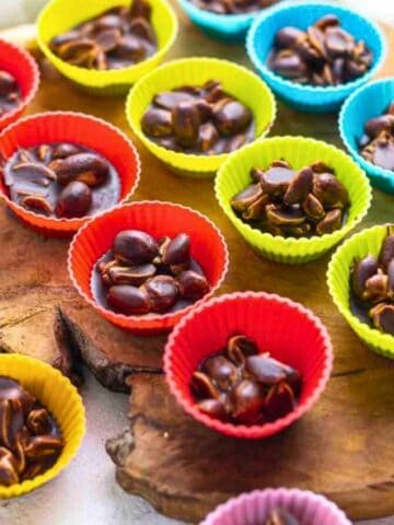 Colorful mini cupcake liners filled with chocolate and peanuts on a wooden board, surrounded by raw peanuts.