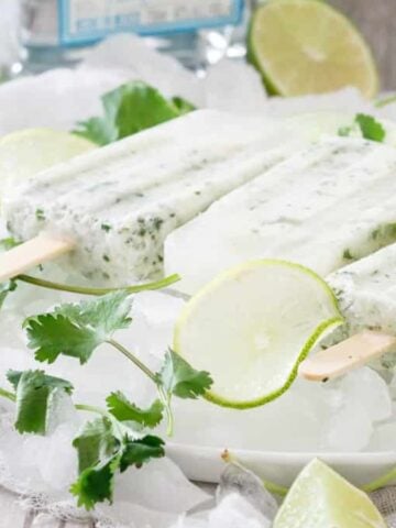 Coconut cilantro margarita ice pops on a plate of ice with fresh cilantro and some slices of lime.