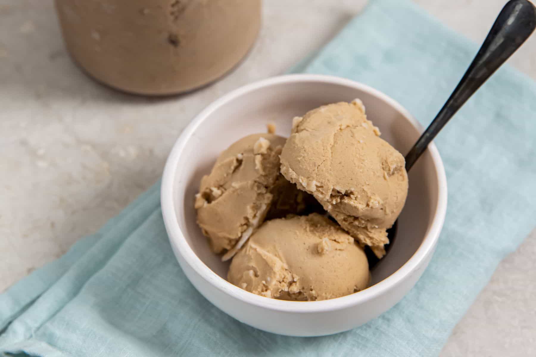 Coffee Ice Cream Ninja Creami in a glass bowl with a spoon and a glass jar with an ice cream scooper.